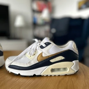 Nike Air Max 90 vit och guld - Nike Air Max 90 sneakers i vit och mörkblå med snygga guldfärgade swoosh-detaljer. Ovandel i mesh och skinn, klassisk Air Max-sula med synlig luftkudde och platt yttersula. Skon har snörning och rund tå, perfekt för dig som gillar retrostil och streetwear. Har använts en gång, så i princip helt nya