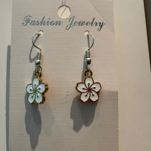 Blommiga örhängen i vitt och guld - Superfina örhängen med blomformade hängen i vitt och guld. Blommorna har detaljerade kronblad och tunna linjer i mitten, vilket ger en söt och fräsch look. Perfekt för dig som vill ha något gulligt och unikt till din stil.