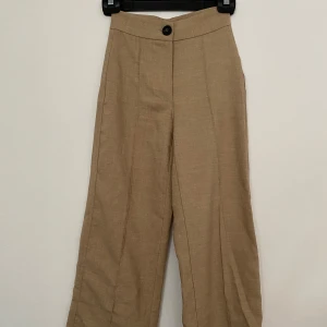 Beiga kostymbyxor från Bershka - Snygga beige byxor från Bershka i storlek 32. Byxorna har hög midja, raka ben och en bred linning med svart knapp framtill. 