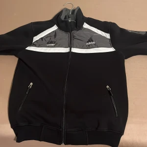 Svart Adidas vindjacka med grå detaljer - Svart vindjacka från Adidas med grå och vita paneler upptill, hel dragkedja och två fickor med dragkedja. Adidas-logga på bröstet och ärmen. Hög krage och sportig look, perfekt för dig som gillar streetwear.