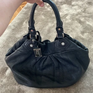 Svart Marc by Marc Jacobs handväska - Säljer snyggaste handväskan från Marc by Marc Jacobs. Väskan har två rejäla handtag, silverfärgade metalldetaljer och en metallbricka framtill. Rymlig modell med snygga rynkade detaljer och coolt innerfoder. Perfekt skolväska 😍