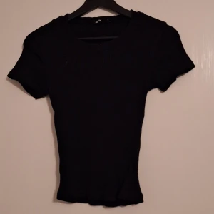 Svart ribbad topp med kort ärm - En svart ribbad topp med rund hals och korta ärmar. Modellen är figurnära och har en enkel, stilren look som passar till det mesta. Materialet är stretchigt och bekvämt, perfekt för dig som gillar en basic men ändå trendig vibe.