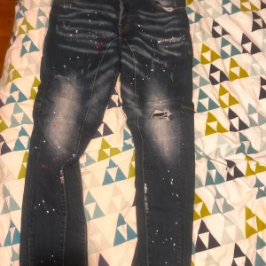 Mörkblå jeans med färgstänk och slitningar - Säljer ett par mörkblå jeans med coola slitningar och färgstänk i vitt och rött. Jeansen har en skinny passform och klassisk femficksdesign. Snygga kontrastsömmar och en unik, streetinspirerad look. Perfekta för dig som gillar en edgy stil. Går perfekt med trade på ett par skor storlek 42 också 
