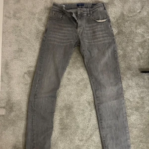 Grå jeans från Scotch & Soda - Säljer ett par grå jeans från Scotch & Soda i klassisk femficksmodell. Jeansen har raka ben och normal passform, tillverkade i bomull med snygga detaljer och diskret tvätt. Perfekta för dig som gillar stilrena och enkla jeans. Storlek 14 år.