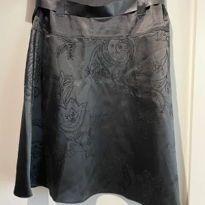 Svart mönstrad satinkjol med rosett - Svart söt satinkjol med subtilt blommönster och paisley-detaljer. Har aldrig används💞