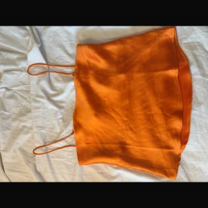 Orange satinlinne med tunna axelband - Snygg orange linnetopp i satinliknande material med tunna axelband och rak halsringning. Toppen har en lätt glansig finish och sitter löst på kroppen för en chill och trendig look. Perfekt att styla med jeans eller kjol för en färgstark outfit.