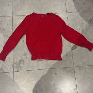 Röd blus med smockade ärmslut - Snygg röd blus från C i storlek 146. Blusen har långärmade smockade ärmslut och ett rynkat band nedtill som ger en cool siluett. Materialet är mjukt och lätt, perfekt för dig som vill ha en färgstark och bekväm topp.