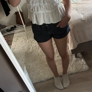 Svarta jeansshorts från Gina Tricot  - Snygga svarta jeansshorts från Gina Tricot med råa kanter och klassisk femficksdesign. Modellen är lågmidjad och har en tight passform som sitter snyggt på kroppen. Perfekta till sommaren och lätta att matcha med allt.