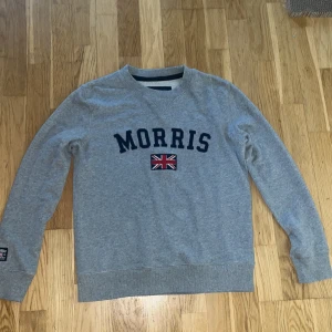 Grå sweatshirt från Morris XS men passar också S - Snygg grå sweatshirt från Morris i storlek XS men passar också S. Tröjan har mörkblå broderad logga och en Union Jack-flagga på bröstet. Rund hals och långa ärmar, mjukt bomullsmaterial som är skönt mot huden. Perfekt för dig som gillar klassisk brittisk stil. Tröjan är i mycket bra skick.