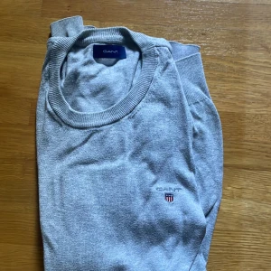 Grå tröja från GANT med broderad logga - Snygg grå tröja från GANT med rund hals och broderad logga på bröstet. Tröjan har ribbade muddar vid hals, ärmslut och nederkant. Perfekt för dig som gillar klassisk och stilren design.