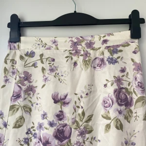 Lång blommig kjol i lila och vitt - Supersöt lång kjol med blommigt mönster i lila och grönt på vit botten. Kjolen har en skön resår i midjan och är gjord i mjuk bomull. Perfekt för dig som gillar romantiska vibes och vill sticka ut med ett färgglatt plagg.