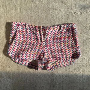 Färgglada mönstrade shorts från ba&sh - Säljer ett par unika shorts från ba&sh med coolt mönster i rosa, blått, gult och rött på vit botten. De har midwaist passform, fickor och blå kantdetaljer. Materialet är grovt och känns lyxigt, perfekt för dig som gillar statement-plagg.