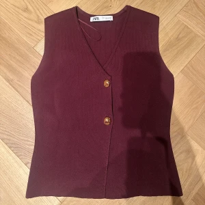 Vinröd stickad väst från Zara - Snygg vinröd stickad väst från Zara med v-ringning och två bruna knappar framtill. Västen har en clean och stilren look, perfekt att styla över en skjorta eller t-shirt. Materialet är mjukt och stretchigt för en skön passform.