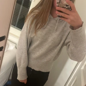Ljusgrå stickad hoodie med huva - Mysig ljusgrå stickad hoodie med huva och snörning. Tröjan har långa ärmar med breda ribbade muddar och en loose passform. Perfekt för chill dagar och när du vill vara extra comfy. Materialet är mjukt och känns skönt mot huden. Använd fåtal gånger och är i bra skick! 