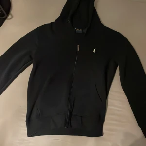 Mörkblå hoodie från Polo Ralph Lauren - Mörkblå hoodie från Polo Ralph Lauren med dragkedja och klassisk vit logga på bröstet. Tröjan har huva med snörning och är tillverkad i mjuk bomullsmix. Perfekt för en chill och stilren look.