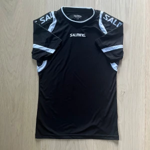 Svart Salming innebandytröja med vita detaljer - Snygg svart innebandytröja från Salming med vita kontrastdetaljer längs axlar och sidor. Tröjan har korta ärmar och rund hals, tillverkad i ett lätt och ventilerande syntetmaterial som passar perfekt för träning och match. Salming-logga på bröst, rygg och axlar.