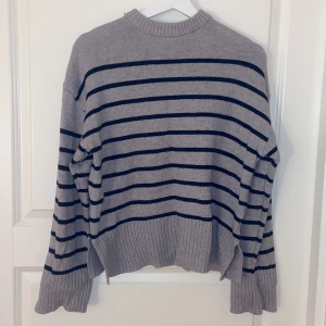 Grå randig stickad tröja med slits - Mysig grå stickad tröja med svarta horisontella ränder. Tröjan har ribbade muddar, högre halsringning och slitsar i sidorna . Den är en Oversized fit med långa ärmar. Och är köpt på lager 157 i storlek S
