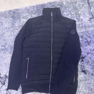 Cardigan Moncler  - Svart dunväst från Moncler med quiltad framsida och stickade ärmar. Västen har hög krage, hel dragkedja och två fickor med dragkedja. Klassisk Moncler-logga på bröstet. Perfekt för lager-på-lager och snygg till streetwear.