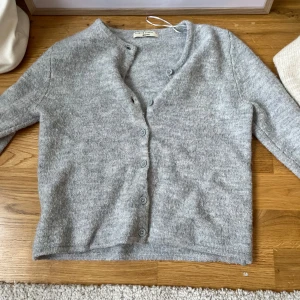 Grå kofta från Gina Tricot, XS - Mysig grå kofta från Gina Tricot i premiumkvalitet. Koftan har v-ringning, långa ärmar och knäppning framtill med matchande knappar. Tillverkad i ett mjukt, stickat material som passar perfekt för lager-på-lager-stil.