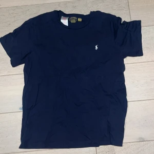 Mörkblå t-shirt från Polo Ralph Lauren - Snygg mörkblå t-shirt från Polo Ralph Lauren med klassisk passform och korta ärmar. T-shirten har en liten vit Polo-logga broderad på bröstet och är gjord i mjuk bomull. Perfekt för en chill och stilren look.