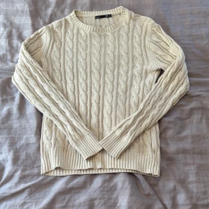 Beige kabelstickad tröja  - Säljer en beige kabelstickad tröja med rund hals och långa ärmar. Tröjan har ett klassiskt flätmönster och ribbade muddar vid ärmslut och nederkant. Perfekt för dig som gillar en chill och stilren look. 