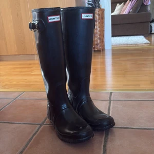  Hunter stövlar - Säljer ett par klassiska svarta Hunter stövlar i gummi med högt skaft och justerbart spänne på sidan. Perfekta för regniga dagar och festivaler. Stövlarna har en rund tå och platt sula, samt den ikoniska Hunter-loggan framtill.