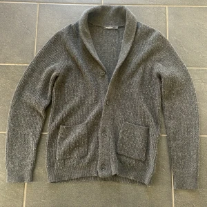 Mörkgrå stickad kofta från Dressmann - Säljer en mörkgrå stickad kofta från Dressmann i storlek M. Koftan har ribbad struktur, två stora fickor framtill och stängs med tre knappar. Perfekt för lager-på-lager och chill stil under höst och vinter.