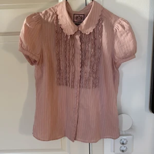 Rosa blus från Juicy Couture - Supersöt ljusrosa blus från Juicy Couture med korta puffärmar, volangdetaljer framtill och rund krage med vågig kant. Skjortan har knäppning framtill. 