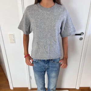 Grå stickad kortärmad tröja - En enkel och stilren grå stickad tröja med korta ärmar och rund halsringning. Tröjan har en rak passform och är gjord i ett mjukt material som känns skönt mot huden. Perfekt för lager-på-lager eller som den är till jeans.