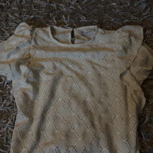 Vit blus med volang från Holly & Whyte - Supersöt vit blus från Holly & Whyte med diskret strukturmönster och volangdetaljer på axlarna. Blusen har rund halsringning och korta ärmar, perfekt för en fräsch och stilren look.