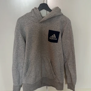 Grå hoodie från Adidas med logga - Grå hoodie från Adidas med klassisk svart logga på bröstet. Tröjan har huva, känguruficka och långa ärmar. Mjuk och skön i bomullsmix, perfekt för chill eller träning. Enkel och stilren design som funkar till det mesta.