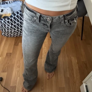 Grå bootcut jeans från True Religion - Säljer ett par gråa bootcut jeans från True Religion med snygga detaljerade bakfickor och silverfärgade knappar. Jeansen har låg midja och klassisk femficksdesign. Perfekta för dig som gillar en avslappnad men ändå trendig look.