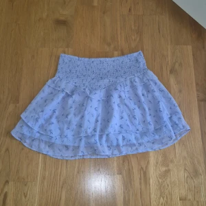 Blommig ljusblå volangkjol Gina Tricot - Supersöt ljusblå kortkjol från Gina Tricot med blommigt mönster och volangdetaljer. Kjolen har en bred, smockad midja som är riktigt bekväm och ger en snygg passform. Perfekt för vår och sommar, och materialet är lätt och luftigt.