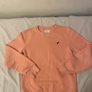 Ljusrosa sweatshirt från Pier One - Säljer en ljusrosa sweatshirt från Pier One med rund hals och långärmad passform. Tröjan har ett litet svart broderat fågelmotiv på bröstet och mjukt fleecefoder på insidan. Perfekt för chill dagar och enkel att matcha med jeans eller joggers.