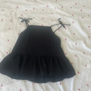 Svart volangtop från H&M med knyt - Supersöt svart linnetopp från H&M med volang nertill och tunna axelband som knyts i rosetter. Perfekt för en chill sommardag eller när du vill känna dig extra söt. Luftig och lätt, passar till både jeans och kjol.