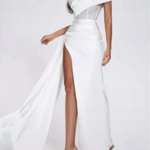 Vit långklänning med slits från Bella Barnett - Supersnygg vit långklänning från Bella Barnett med glittrande detaljer på överdelen och en dramatisk hög slits. Klänningen har omlott-effekt och är axelbandslös med ett lyxigt satinliknande material som ger en elegant vibe. Perfekt för bal eller fest!