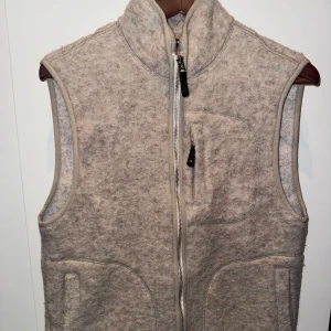 Beige ullväst från J.Lindeberg - Snygg beige väst från J.Lindeberg i mjuk ullmix. Västen har hög krage, hel dragkedja framtill och praktiska fickor, inklusive en bröstficka med zip. Perfekt att slänga över en hoodie eller långärmad för extra värme. Stilren och enkel design.