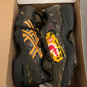ASICS x BAPE camo sneakers strl 44 - Unika ASICS x BAPE sneakers med camouflage-mönster, orangea detaljer och ikonisk tiger-grafik på sidan. Skorna har svarta och gröna toner, snörning och robust svart sula. Tillverkade i syntet och mocka med gummisula. Perfekta för dig som vill sticka ut.