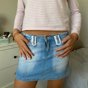 Sjukt snygg blå jeanskjol - Säljer en kort jeanskjol i ljusblå tvätt med slitna detaljer och broderad text 'New Crazy' på linningen bak. Kjolen har fram- och bakfickor, bälteshällor och knappstängning. Perfekt för dig som gillar streetstyle och denim.