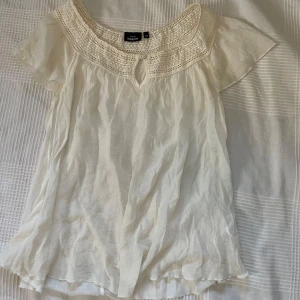 Vit blus med spets från KappAhl XS - Supersöt vit blus från KappAhl i storlek XS. Blusen har korta ärmar, rund halsringning med virkade spetsdetaljer och en liten öppning framtill. Luftigt material.