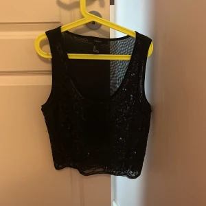 Svart paljettopp från Forever 21 - Säljer en svart topp från Forever 21, täckt med glittriga paljetter framtill. Toppen är ärmlös och har en transparent mesh-baksida som ger en snygg kontrast. Perfekt för dig som vill sticka ut med lite extra bling. 