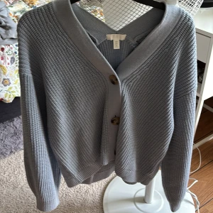 Ljusblå stickad kofta från H&M - Säljer en ljusblå stickad kofta från H&M i storlek M. Koftan har grov ribbad struktur, två stora bruna knappar framtill och en djup v-ringning. Långärmad och med breda muddar vid ärmslut och nederkant. Perfekt för lager-på-lager under kyliga dagar.