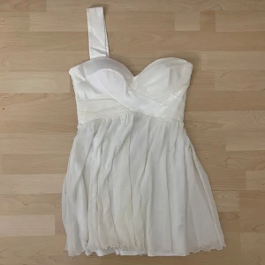 Vit axelbandslös kort klänning - Säljer en vit kort klänning med ett axelband och sweetheart-urringning. Klänningen har omlott-detaljer upptill och ett lätt, flödande tyg nedtill som ger en romantisk vibe. Perfekt för student eller sommarens alla tillfällen!