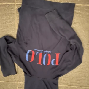 Mörkblå hoodie från Polo Ralph Lauren - Säljer en mörkblå hoodie från Polo Ralph Lauren med stor POLO-logga i rött och blått på ryggen och liten broderad logga på bröstet. Tröjan har dragkedja framtill och huva. Perfekt för en avslappnad stil.