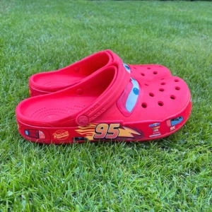 Röda Crocs med Blixten McQueen motiv - OBS!! Bandens har tyvärr försvunnit på crocsen men är fortfarande lika snygga och snabba Säljer ett par röda Crocs med Blixten McQueen från Disney Cars. Skorna har öppen tå, platt sula och coola detaljer som ögon och nummer 95 på sidorna. Perfekta för dig som gillar färgglada och lekfulla skor!