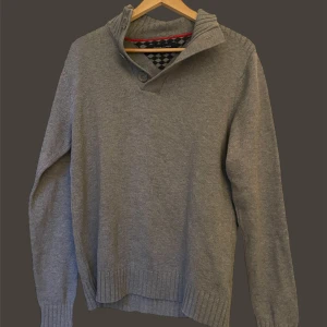 Tommy hilfiger tröja  - Säljer en grå stickad tröja med hög krage och två knappar vid halsen. Tröjan har långa ärmar och ribbade muddar vid ärmslut och nederkant. Perfekt för kyliga dagar!