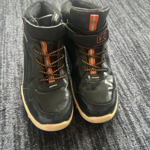 Svarta höga sneakers från Friboo med orange detaljer - Säljer ett par svarta höga sneakers från Friboo med orangea och vita detaljer. Skorna har snörning, kardborreband och vit sula. Insidan är fodrad med mjukt tyg för extra komfort. Perfekta för dig som gillar sportig stil.