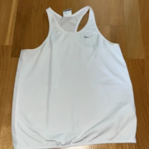 Vit träningstopp från Nike - Säljer en vit linnetopp från Nike med brottarrygg och diskret Nike-logga framtill. Perfekt för träning och sportiga aktiviteter. Tillverkad i ett lätt och syntetiskt material som andas. Är barnstorlek L men är som en xs.