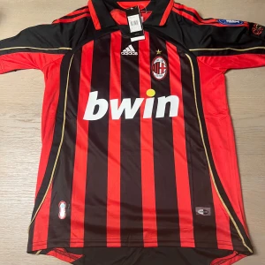 AC Milan fotbollströja Kaka #22 - Säljer en klassisk AC Milan fotbollströja med Kaka och nummer 22 på ryggen. Tröjan är svart och röd med vertikala ränder, korta ärmar och guldiga detaljer. Adidas-logga och Bwin som sponsor på framsidan.