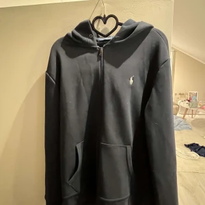 Svart hoodie från Polo Ralph Lauren - Säljer en svart hoodie från Polo Ralph Lauren med dragkedja och broderad logga på bröstet. Tröjan har känguruficka och huva i en något mörkare nyans. Perfekt för en avslappnad stil. Nästan helt oanvänd. Ny pris 1500 kr, pris kan diskuteras 